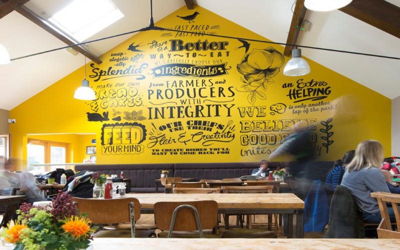 Image of our cafe. Wooden topped tables and chairs. Gabled roof with wooden beams. 
                Large yellow feature wall with key words written in a range of interesting font styles.