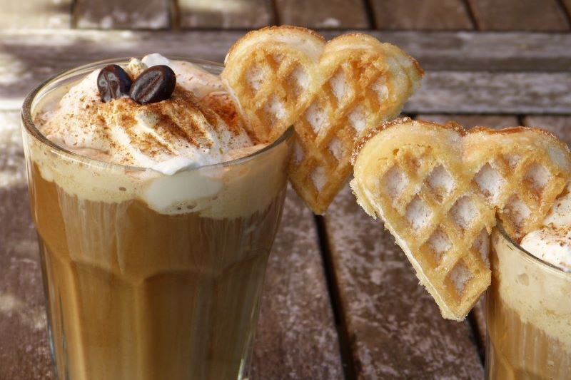 Tall glass of ice coffee with cream on top and hart shaped waffle biscuit on edge of glace.